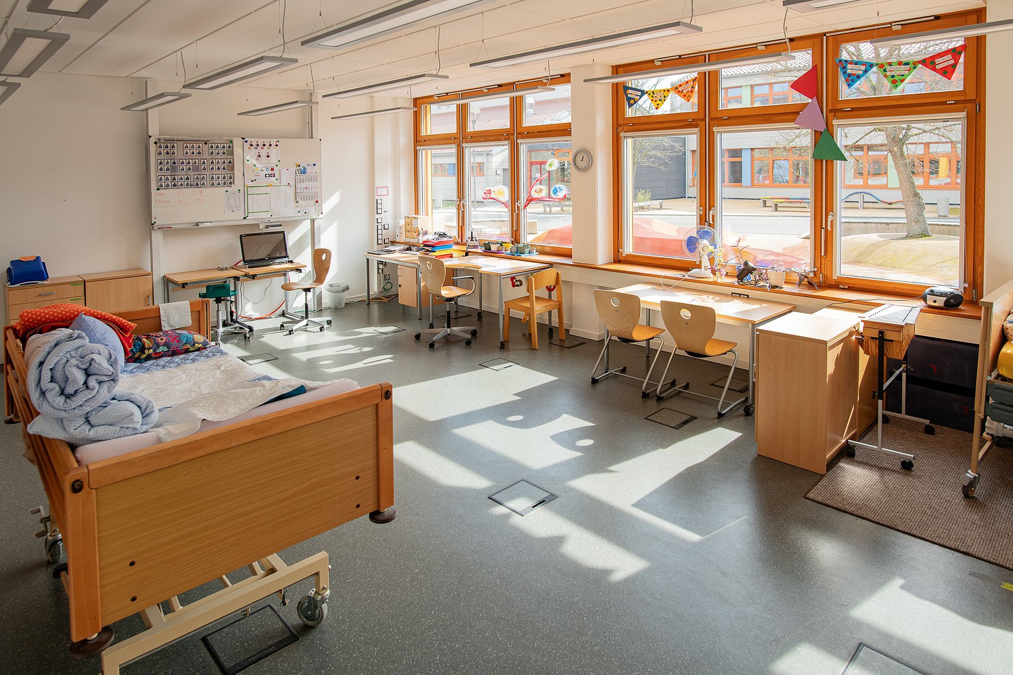 Ein Klassenzimmer, in dem rechts an der Fensterfront Schreibtische und Stühle stehen. Vorne an der Wand hängt eine Tafel. Im linken Teil steht ein Pflegebett. Das Klassenzimmer ist sehr hell und mit warmen Farben gestaltet. 