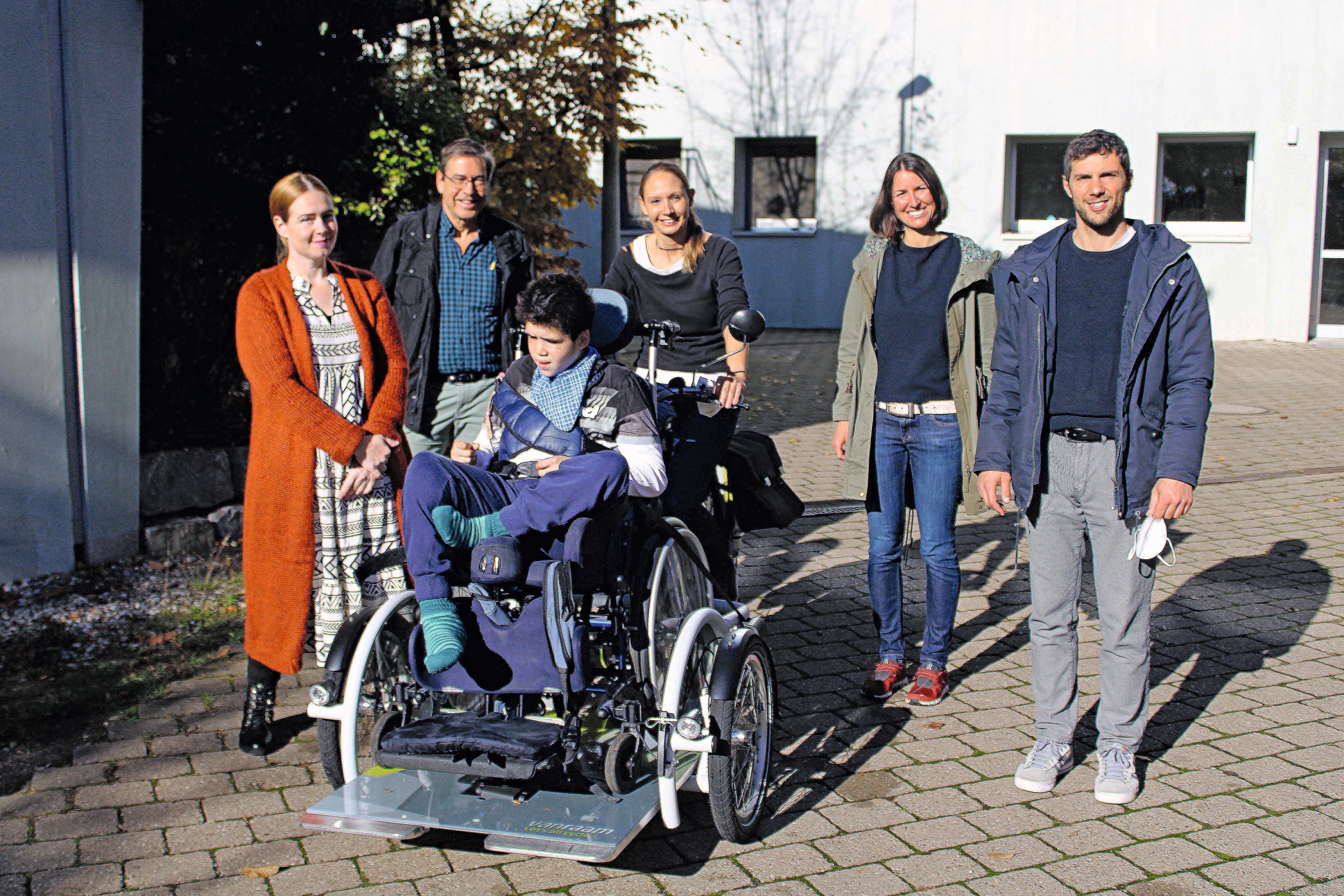 Ein Junge auf einem Rollstuhl-Fahrrad. Dahinter stehen fünf Personen um den Jungen verteilt. 