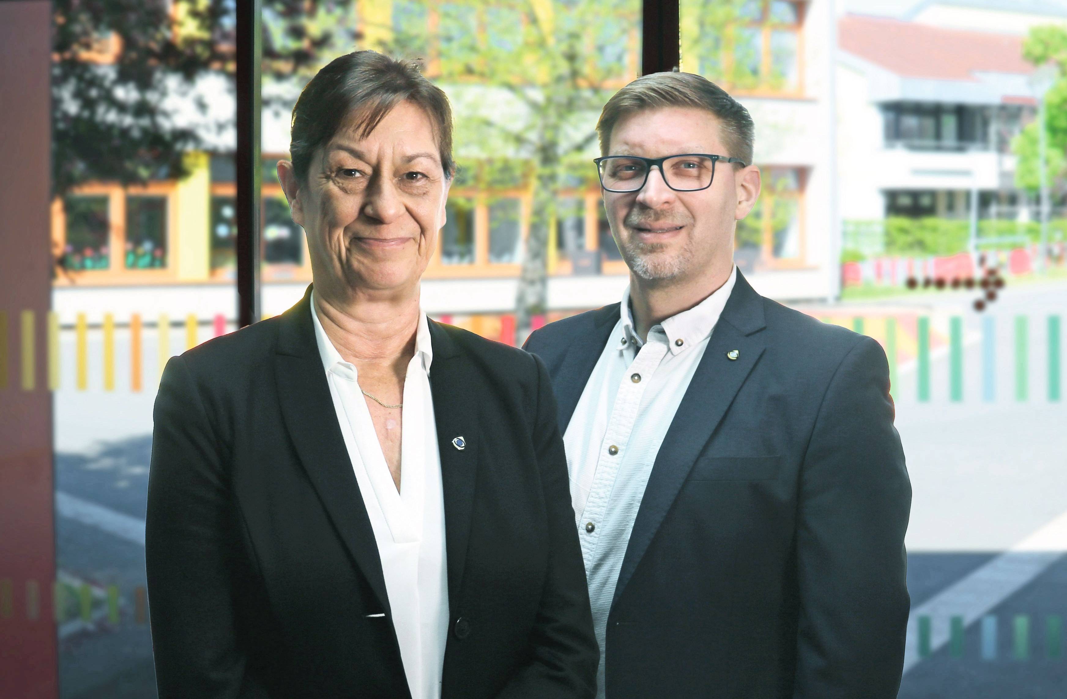 Heike Sandrock und Frank Tollkühn. Beide haben dunkle Anzüge an. Frau Sandrock steht links vor Herrn Tollkühn. 