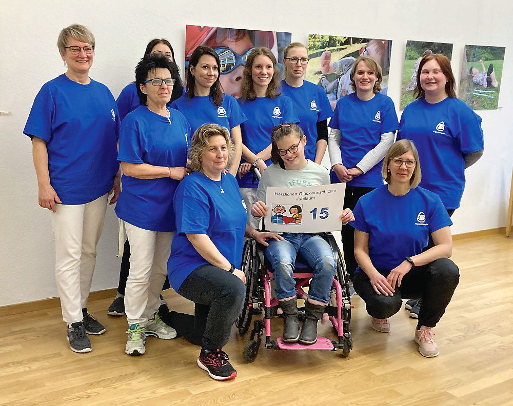 Im Fokus ist ein Mädchen in einem rosa Rollstuhl. Sie hält ein Schild in der Hand. Auf diesem steht „Herzlichen Glückwunsch zum Jubiläum – 15“. Um sie herum stehen 10 Frauen, die alle blaue T-Shirts tragen, auf dem das Blindeninstitutslogo zu sehen ist. 