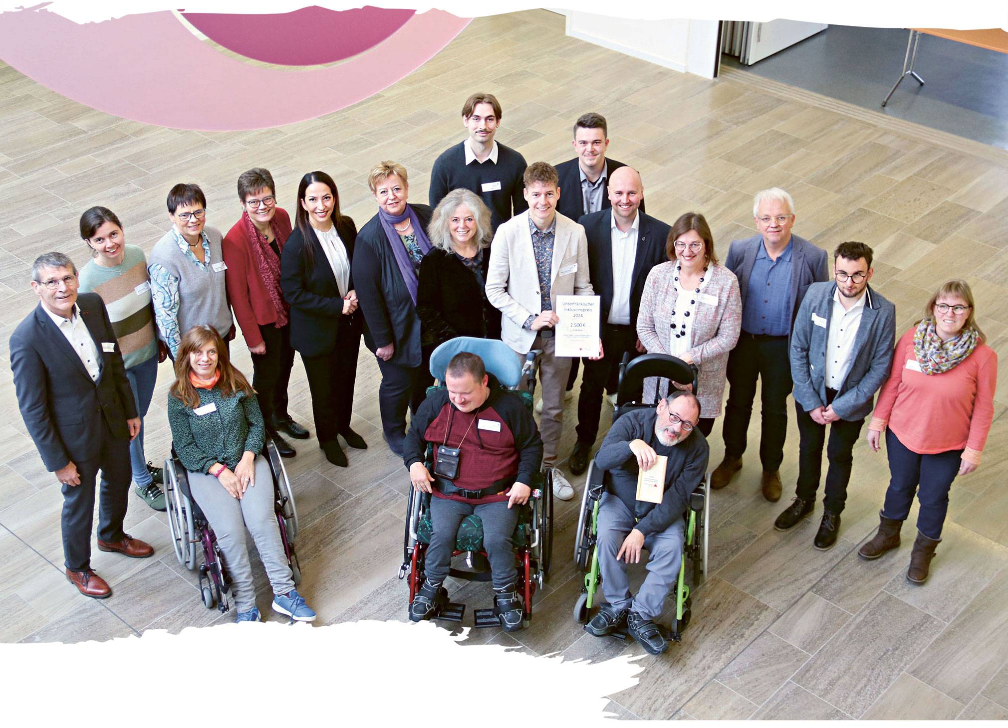Eine Gruppe von 18 Personen steht in einem offenen, modernen Innenraum zusammen. Drei Personen sitzen im Rollstuhl, die anderen stehen. In der Mitte hält eine Person den Inklusionspreis in Form einer Urkunde in der Hand.