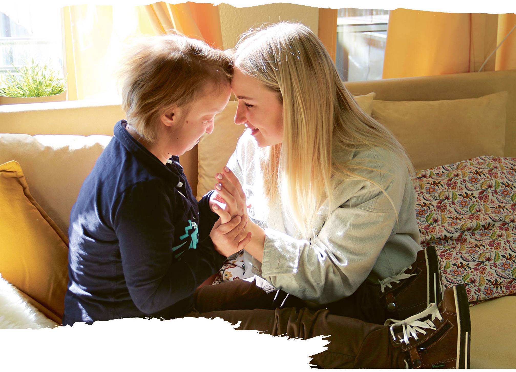 Zwei Personen, ein Kind und eine junge Frau mit langen blonden Haaren, sitzen sich auf einem Sofa gegenüber. Sie berühren liebevoll ihre Stirnen und lachen miteinander in einer hellen, gemütlichen Umgebung mit gelben Vorhängen.