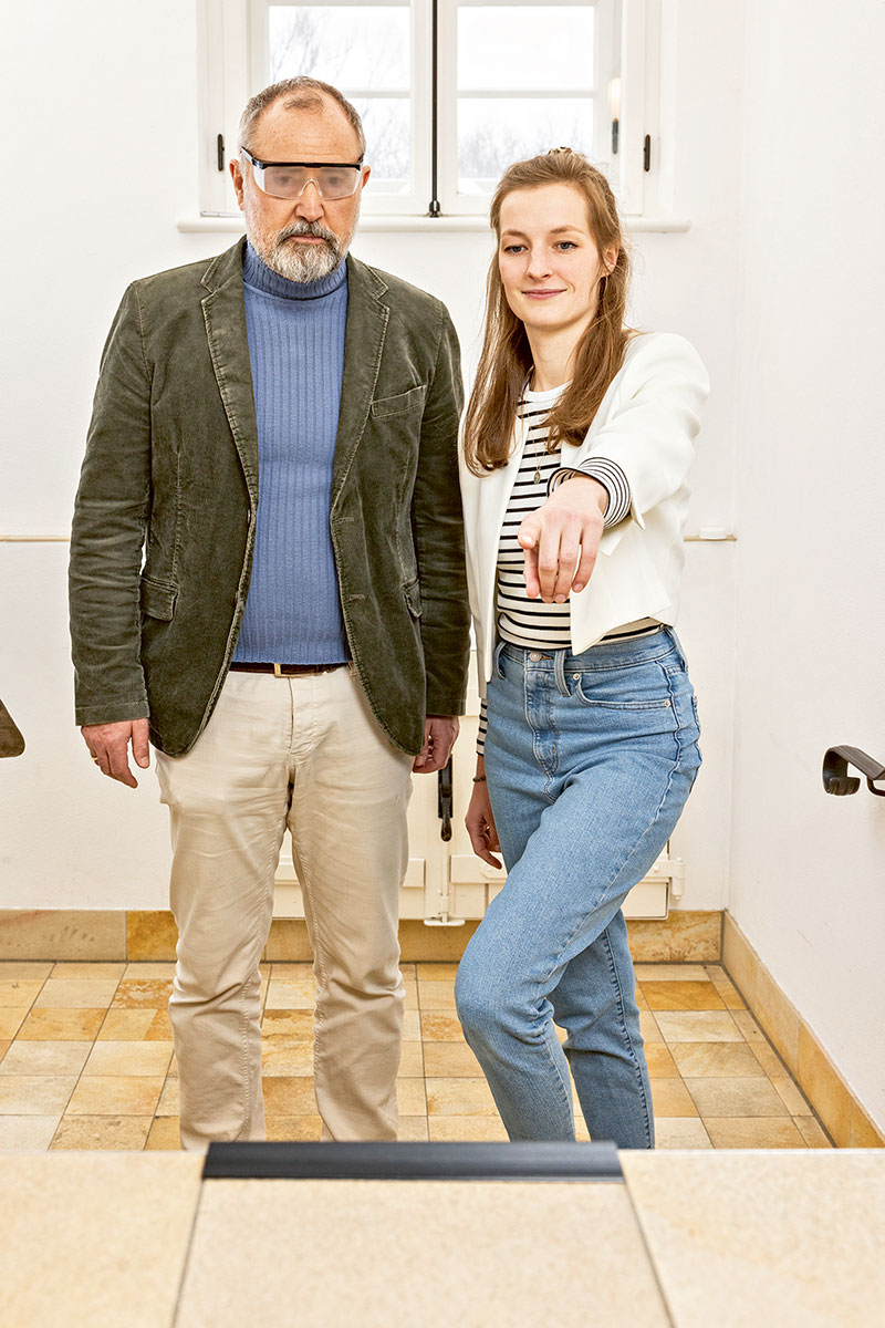 Ein Mann mit Simulationsbrille steht neben einer Frau, die auf etwas vor ihnen zeigt. Sie befinden sich in einem hellen Raum mit Fliesenboden.