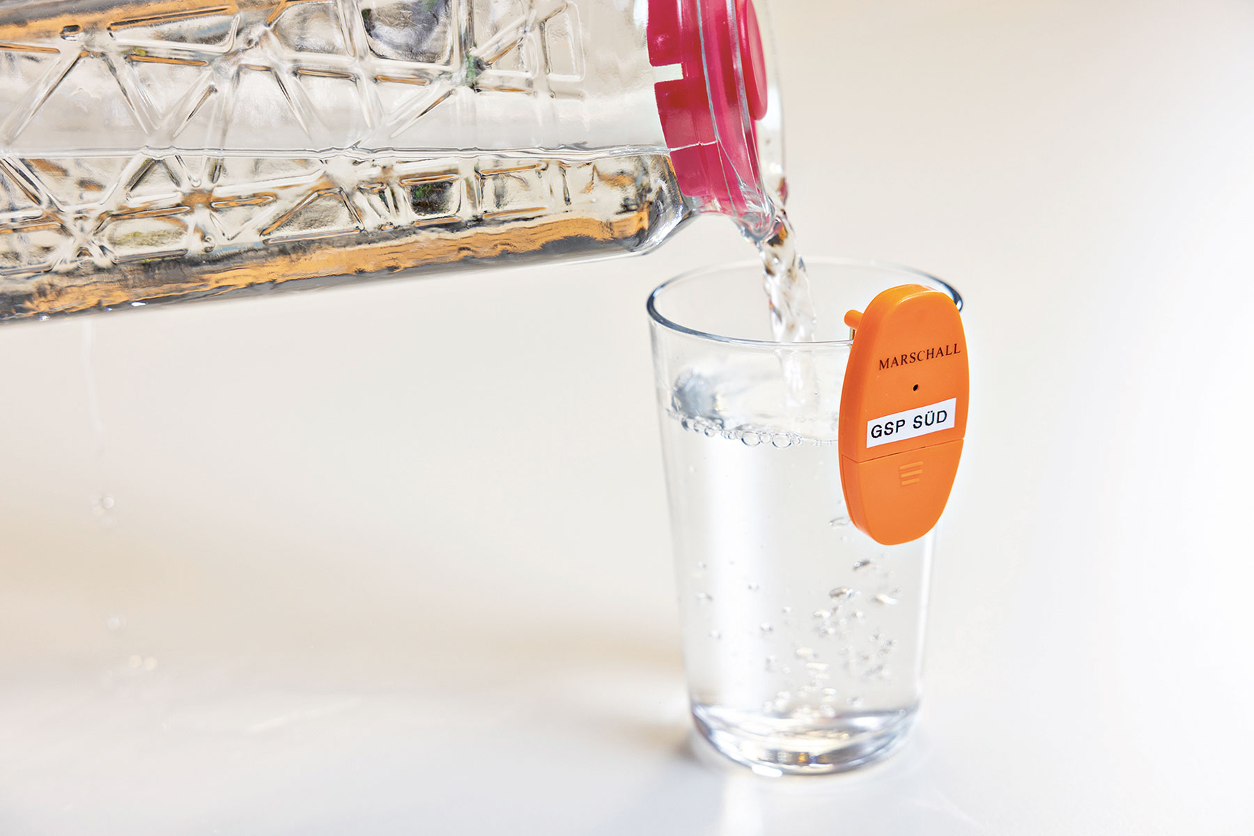 Ein Glas wird mit Wasser aus einer Karaffe gefüllt. Am Glas hängt ein orangefarbener Füllstandsanzeiger.