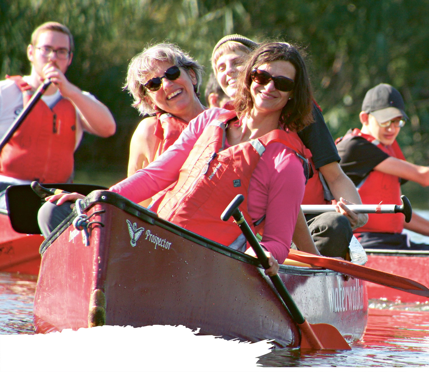 Schüler und Betreuer in roten Schwimmwesten sitzen lachend in drei roten Kanus auf dem Wasser. Sie paddeln gemeinsam und wirken fröhlich. Im Hintergrund sind grüne Pflanzen am Ufer zu sehen.