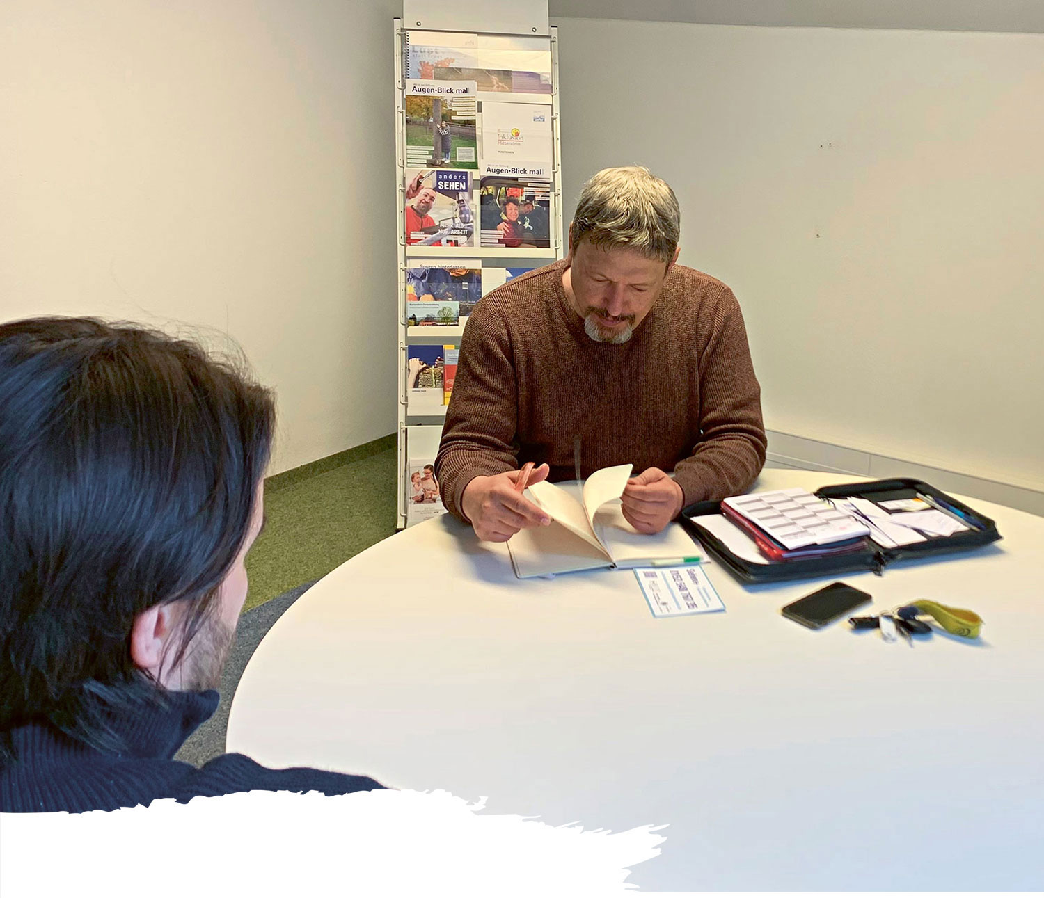 Zwei Personen sitzen sich an einem runden Tisch gegenüber. Die eine Person blättert in einem Heft, vor ihr liegen Schreibutensilien und ein Etui. Die andere Person ist von hinten zu sehen. Im Hintergrund steht ein Prospektständer.