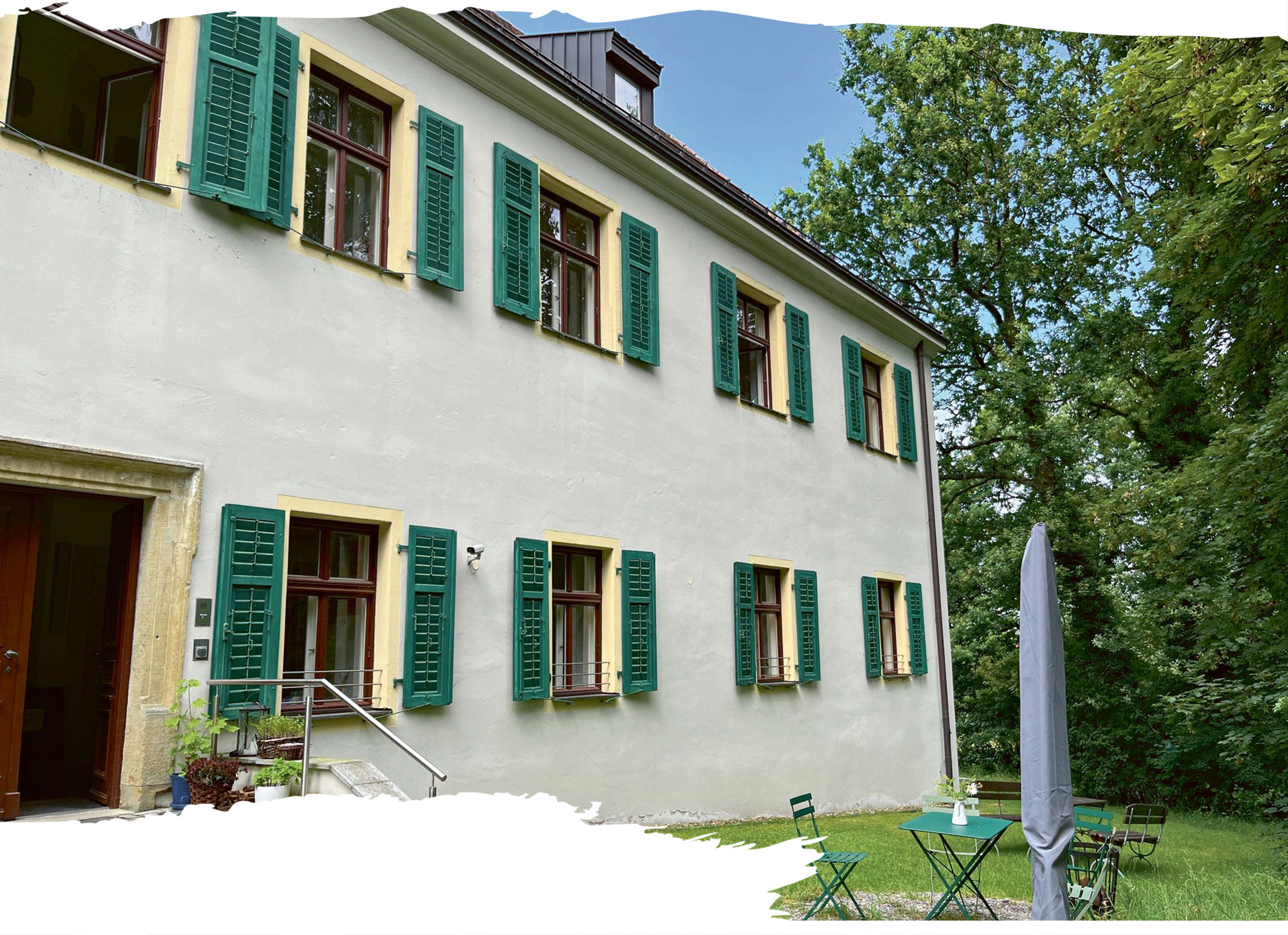 Zweistöckiges, helles Gebäude mit grünen Fensterläden. Davor ein kleiner Tisch mit zwei grünen Gartenstühlen und ein geschlossener Sonnenschirm. Im Hintergrund ein dichter Baum.