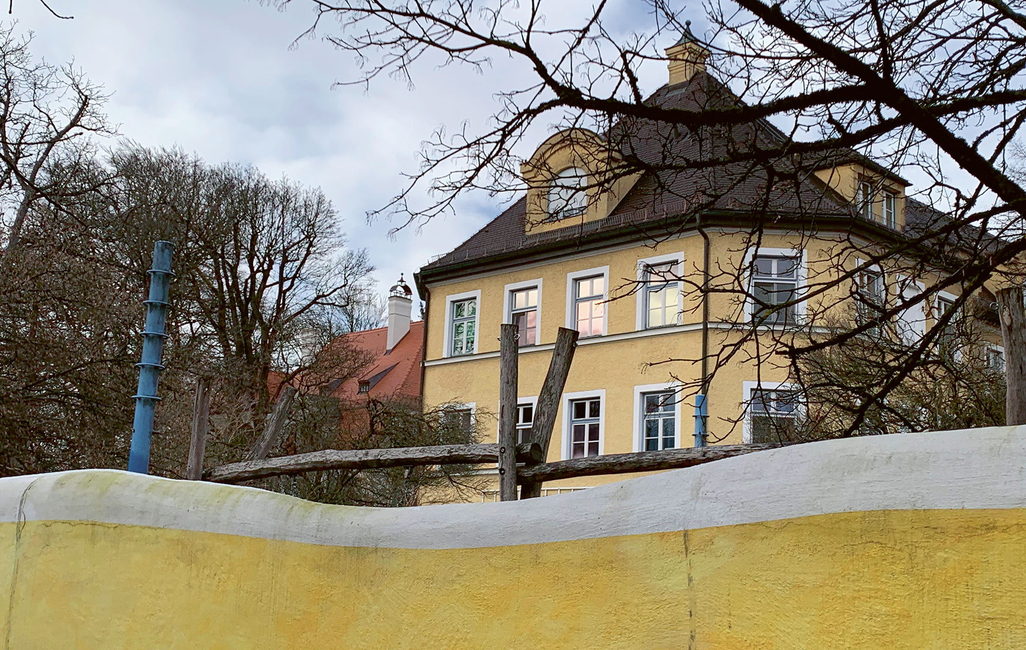 Ein gelbes, mehrstöckiges Gebäude mit dunklem Dach und Dachgauben steht hinter einer geschwungenen gelben Mauer. Im Vordergrund sind kahle Äste zu sehen, im Hintergrund weitere Gebäude und Bäume. Der Himmel ist bewölkt.