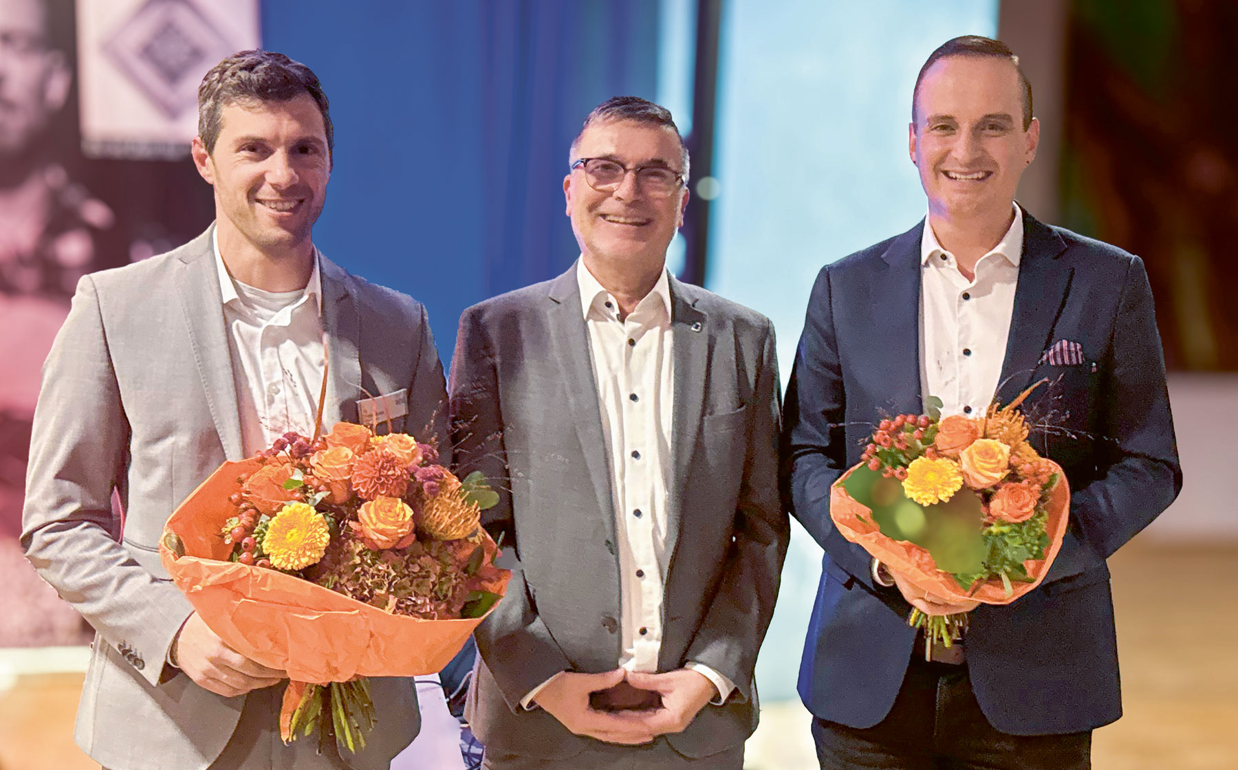 Von links nach rechts: Dominic Weufen, Johannes Spielmann und Daniel Boldt stehen nebeneinander und lächeln in die Kamera. Dominic Weufen und Daniel Boldt halten jeweils einen Blumenstrauß mit orangefarbenen und gelben Blumen in der Hand.