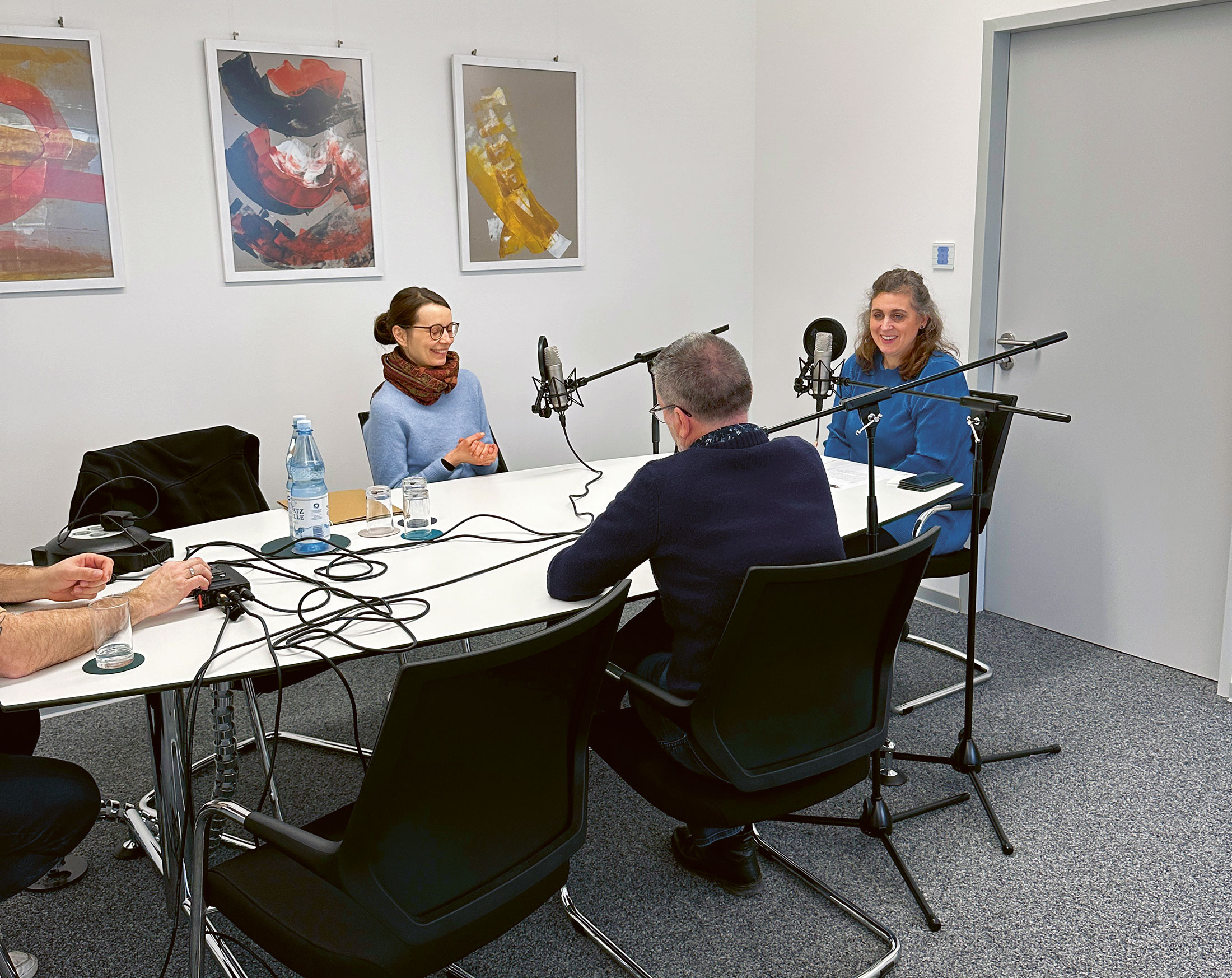 Vier Personen sitzen um einen Tisch und nehmen ein Interview auf. Auf dem Tisch stehen Mikrofone, Kopfhörer und Wasserflaschen. Im Hintergrund eine geschlossene Tür und drei bunte Bilder an einer weißen Wand.