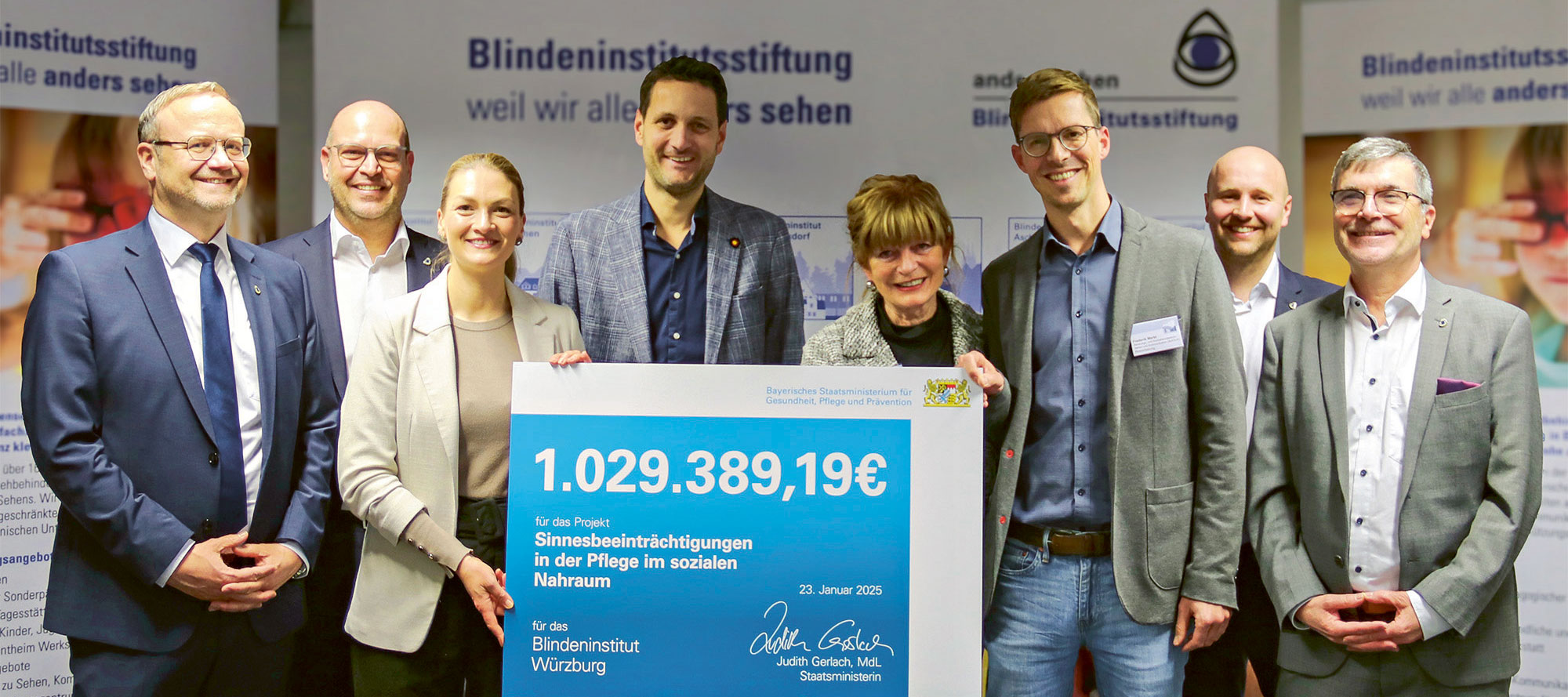 Gruppenfoto mit acht Personen im Blindeninstitut Würzburg. Sie halten gemeinsam einen großen symbolischen Förderscheck in Höhe von 1.029.389,19 Euro. Im Hintergrund sind Roll-Ups der Blindeninstitutsstiftung zu sehen.
