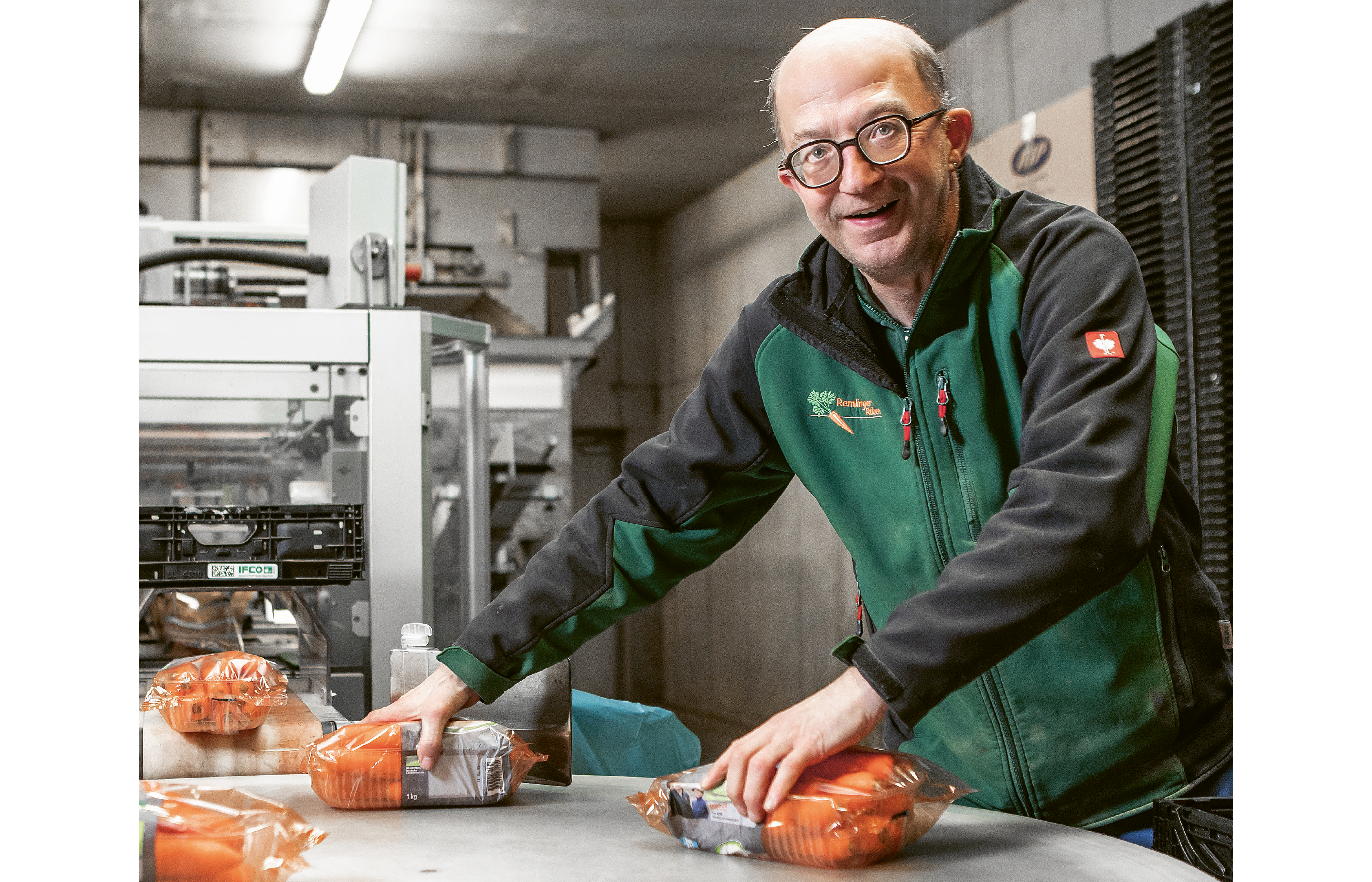 Michael Riethmann bei seiner Arbeit auf dem Rübenhof in Remlingen. Er hat zwei verpackte Schalen Karotten in der Hand, die er aus einer Maschine in eine Kiste verlädt. Er lacht in die Kamera.
