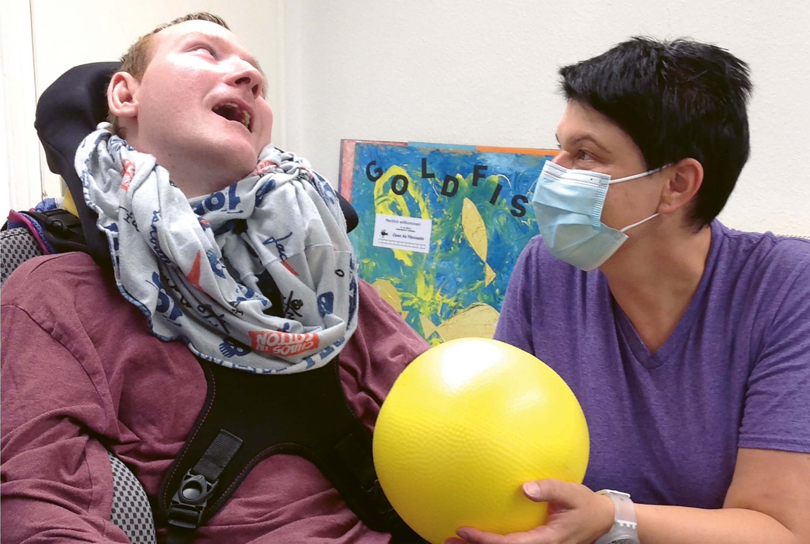 Steffi Kallenbach, die einen Mundschutz trägt, kniet neben einer Person im Rollstuhl und hat einen gelben Ball in der Hand.