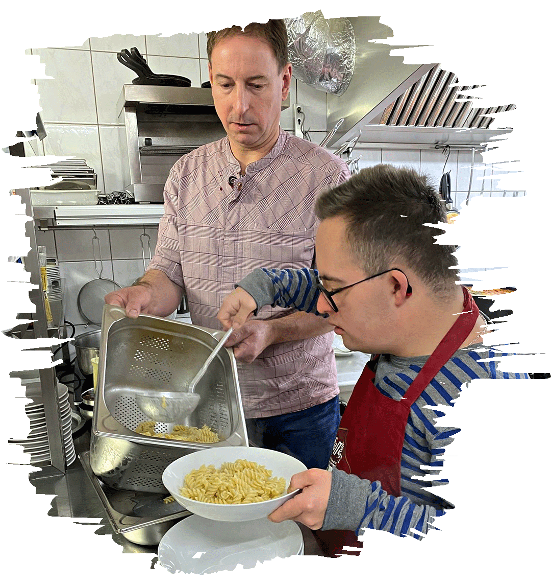 Ein Lehrer und ein Schüler in einer Küche bereiten gemeinsam Essen zu. Der Lehrer hält ein Sieb mit gekochten Nudeln, während der Schüler die Nudeln in einen Teller füllt. Im Hintergrund sind Küchenutensilien und Regale mit Geschirr zu sehen.