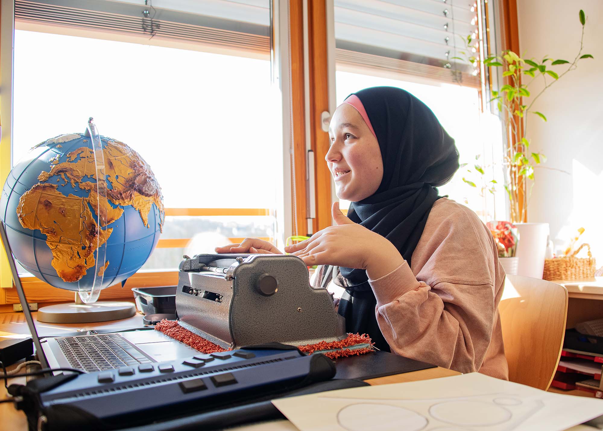 Eine junge Frau mit schwarzem Kopftuch sitzt an einem Schreibtisch und tippt auf einer Braille-Schreibmaschine. 