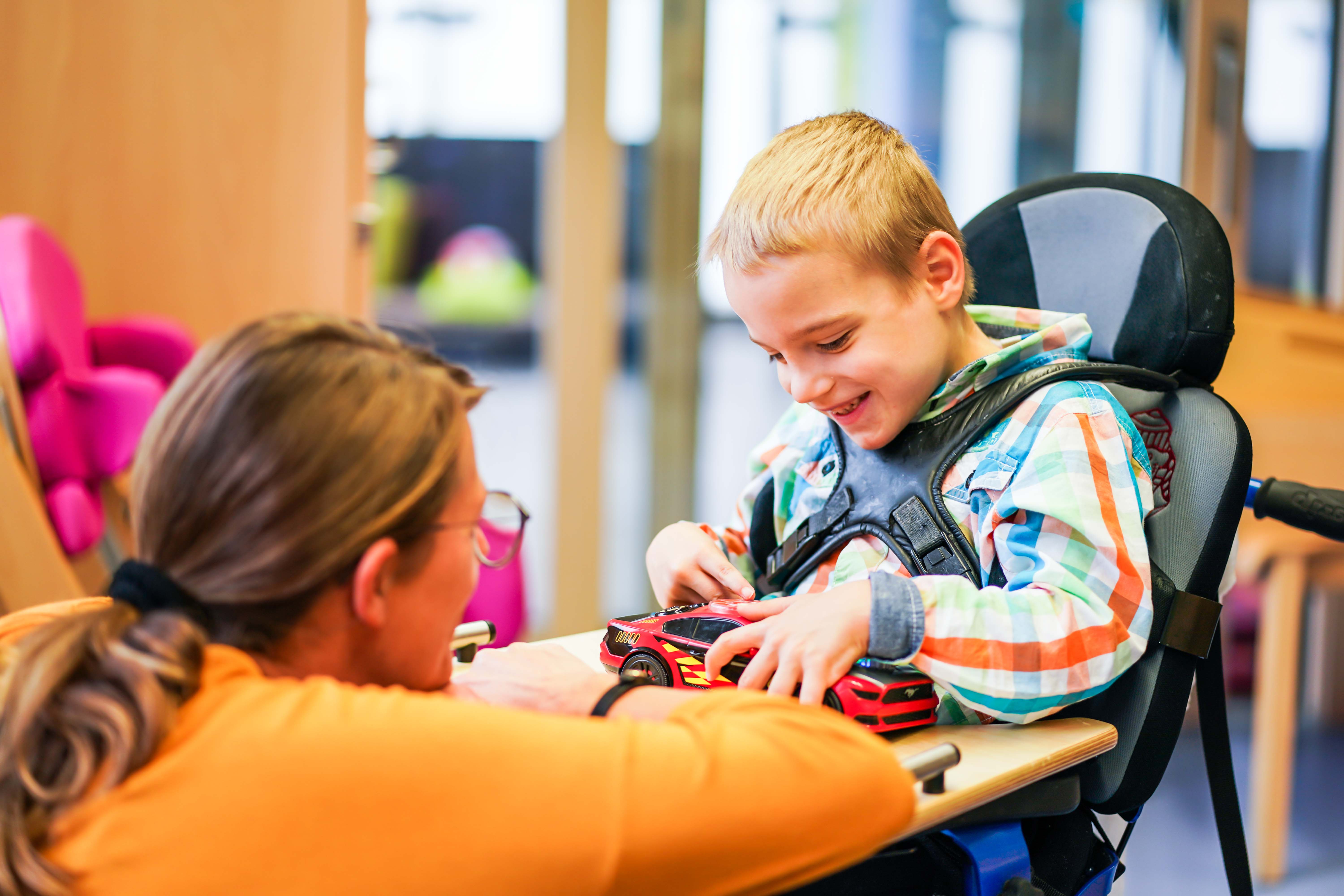 Ein kleiner Junge sitzt in einem Rollstuhl und hält lächelnd ein  Spielzeugauto in der Hand. Vor ihm kniet eine Frau und schaut ihn an.